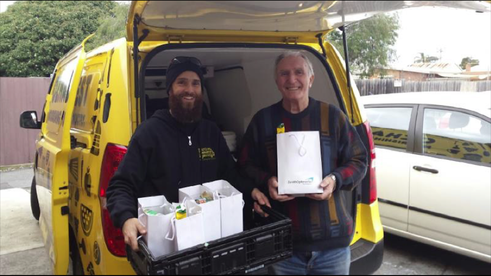 Two men stand by a van holding food parcels, smiling. 